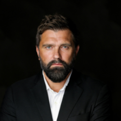 Älterer Mann mit grauen Haaren trägt weißen Hemd und dunklen Anzug und lächelt im Freien.