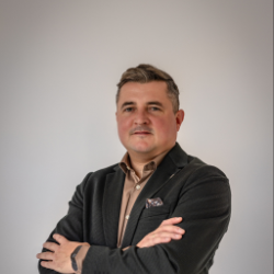 Mann mit verschränkten Armen, trägt schwarzen Blazer und helles Hemd, vor hellem Hintergrund.