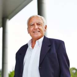 Älterer Mann in weißem Hemd und dunkelblauem Sakko lächelnd vor hellem Hintergrund.