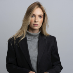 Porträt eines Mannes mit verschränkten Armen, der eine schwarze Strickjacke und ein hellbraunes Hemd trägt.