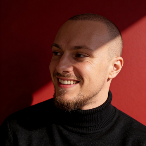 Junger Mann mit lockigem blonden Haar, weißem Hemd und dunklem Sakko vor schwarzem Hintergrund.