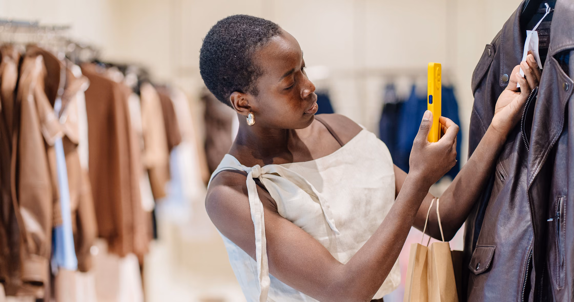 Woman in a store scans the price tag of a brown leather jacket with a yellow smartphone.