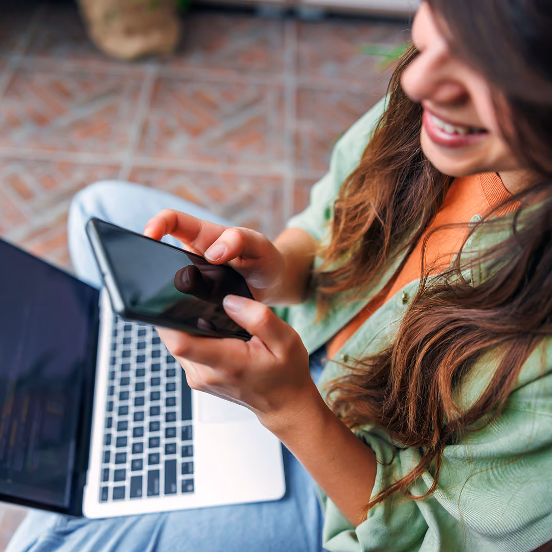 Smiling woman holding smartphone with laptop on her lap, sitting on tiled floor.