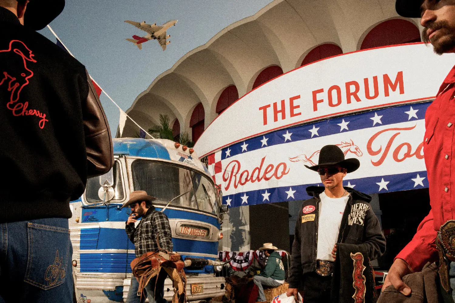 Eine Gruppe von Männern in Cowboy-Kleidung steht vor einem Gebäude, das mit "THE FORUM Rodeo Today" beschriftet ist. Im Hintergrund ist ein Bus mit der Aufschrift "Los Angeles" zu sehen und am Himmel fliegt ein Flugzeug.