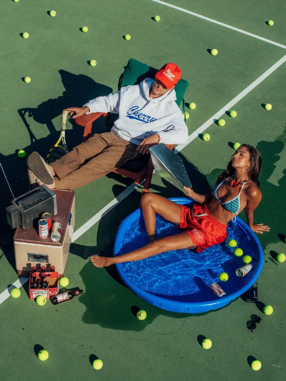Eine Aufnahme von oben, die zwei Personen auf einem Tennisplatz zeigt. Ein Mann sitzt in einem Stuhl mit einer Tennistasche daneben, während eine Frau in einem aufblasbaren Pool liegt. Überall auf dem Platz sind Tennisbälle verstreut.