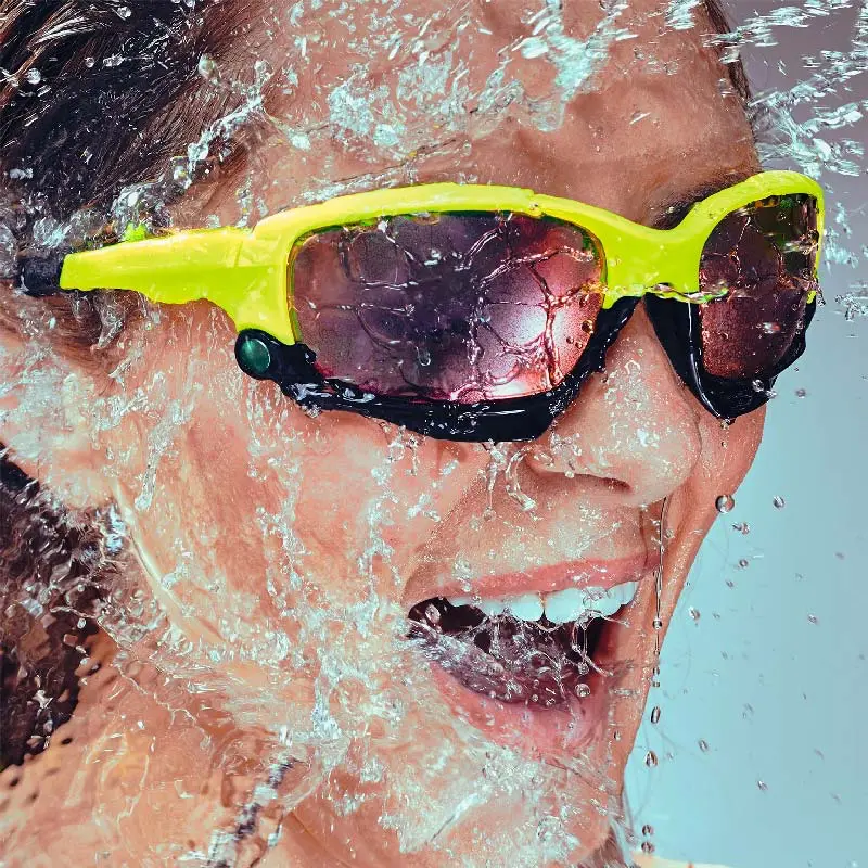 Eine Nahaufnahme eines Gesichts, das eine gelbe Sonnenbrille trägt. Wasser spritzt über das Gesicht und die Sonnenbrille.