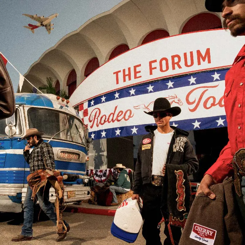 Eine Gruppe von Männern in Cowboy-Kleidung steht vor einem Gebäude, das mit "THE FORUM Rodeo Today" beschriftet ist. Im Hintergrund ist ein blauer Bus mit der Aufschrift "Los Angeles" zu sehen.