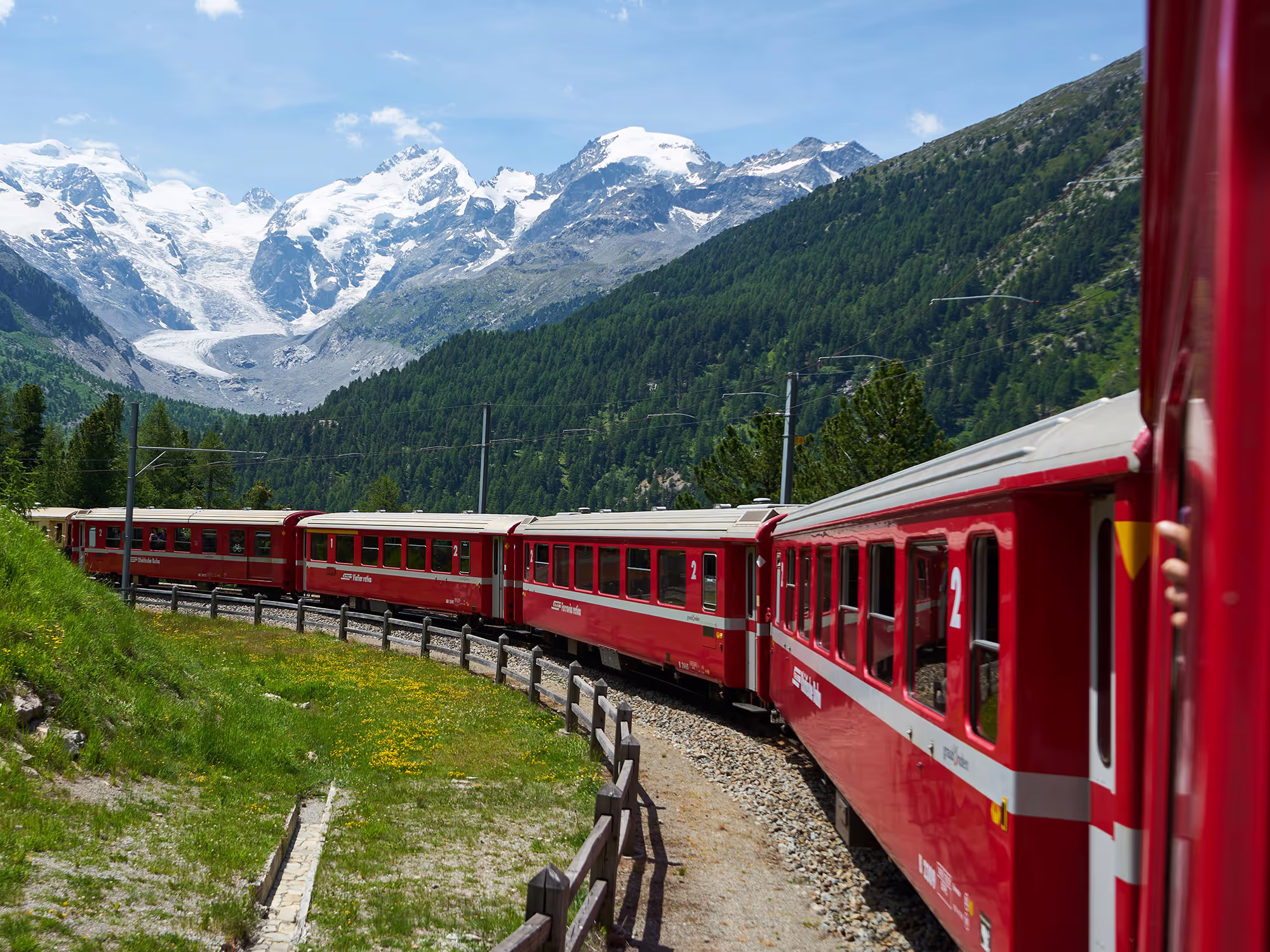 A train navigates a sharp left turn in the Swiss mountains, winding through a breathtaking landscape of steep slopes and lush greenery
