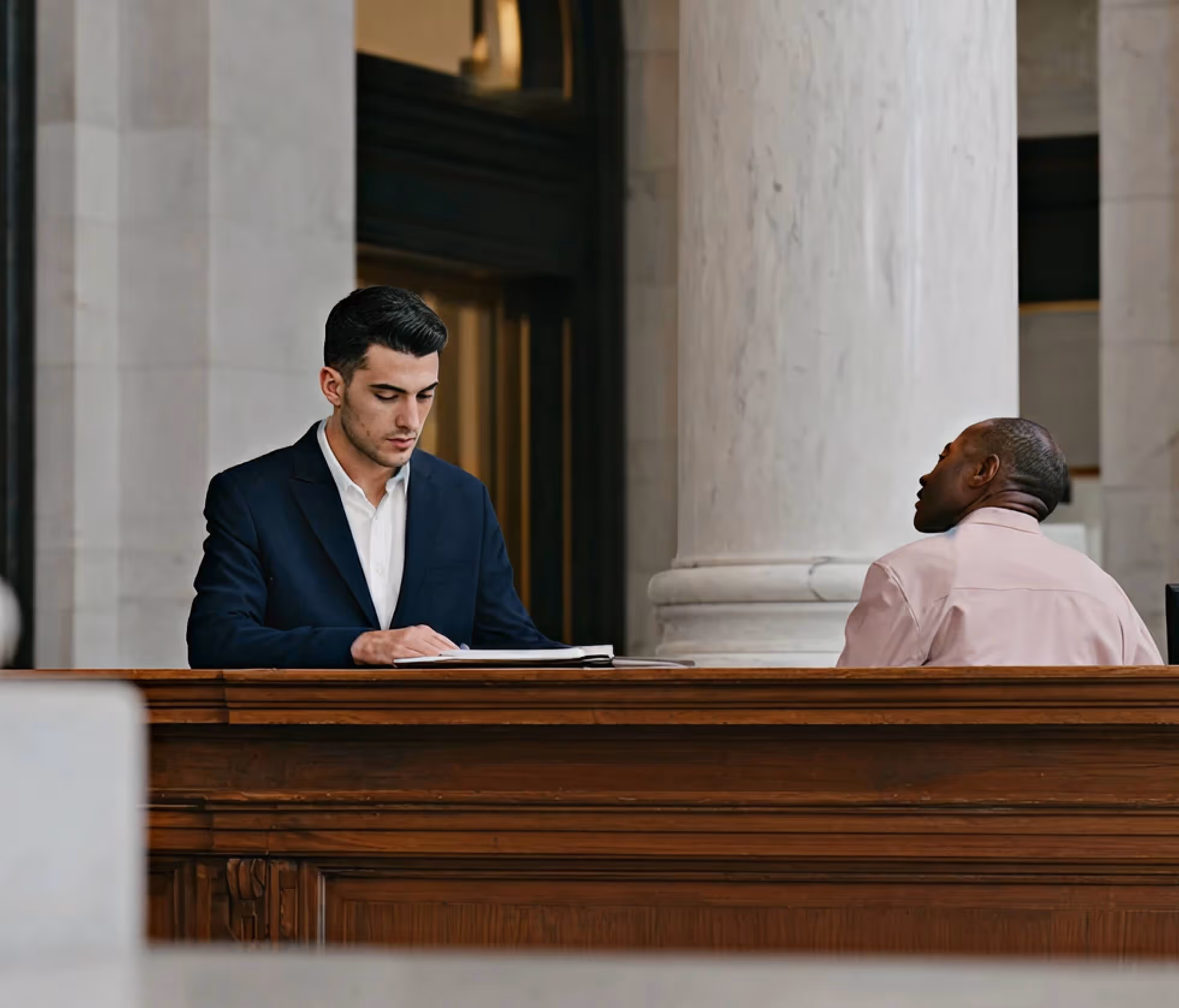 Two men seated at a wooden desk; one reads a document, the other looks to the right in a formal setting.
