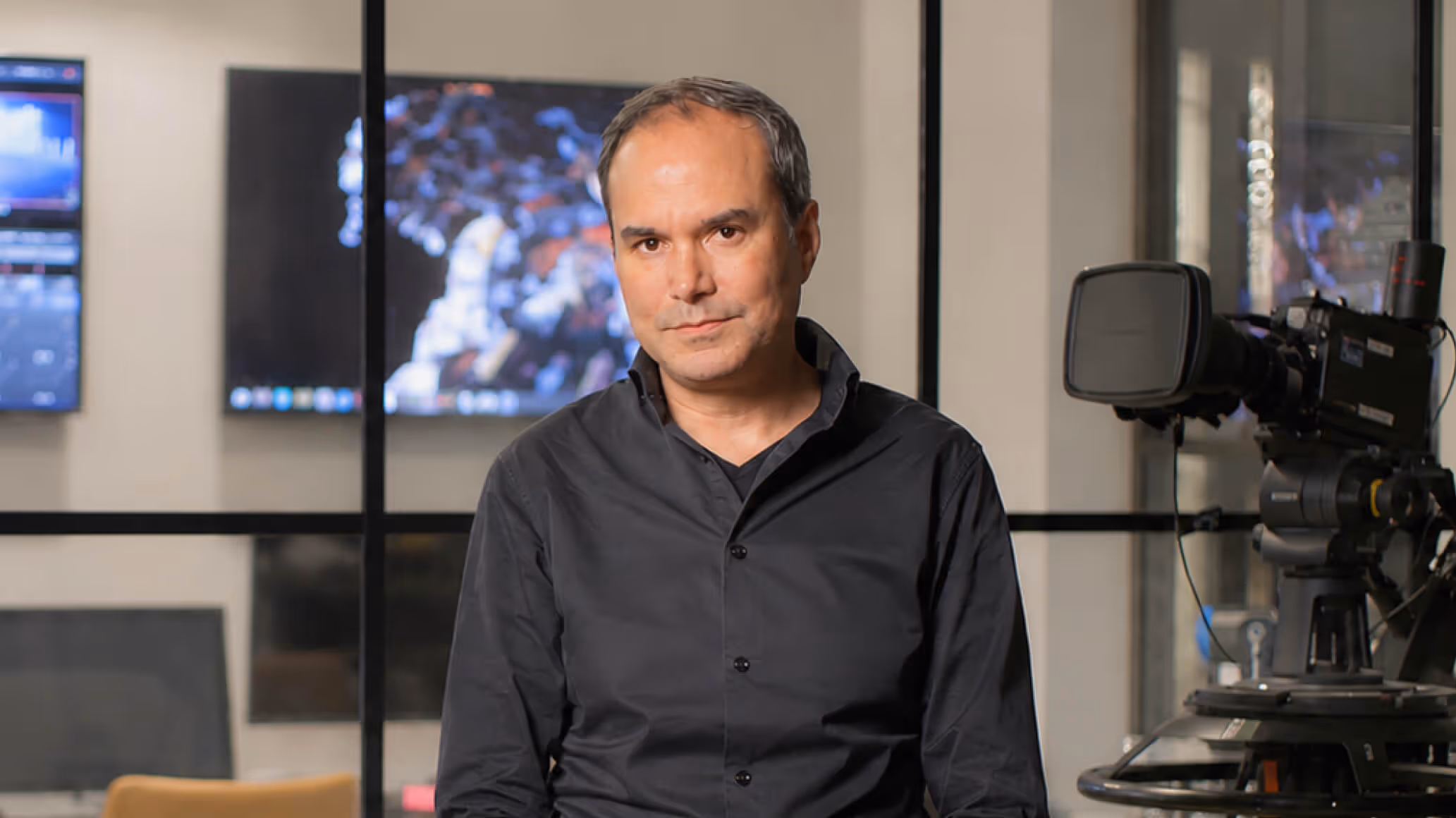 Man in a black shirt sitting in a studio with a professional video camera and screens in the background.