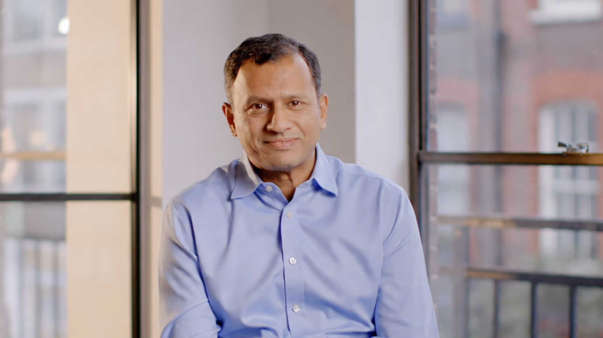 Man in a light blue shirt sitting and smiling indoors with large windows in the background.