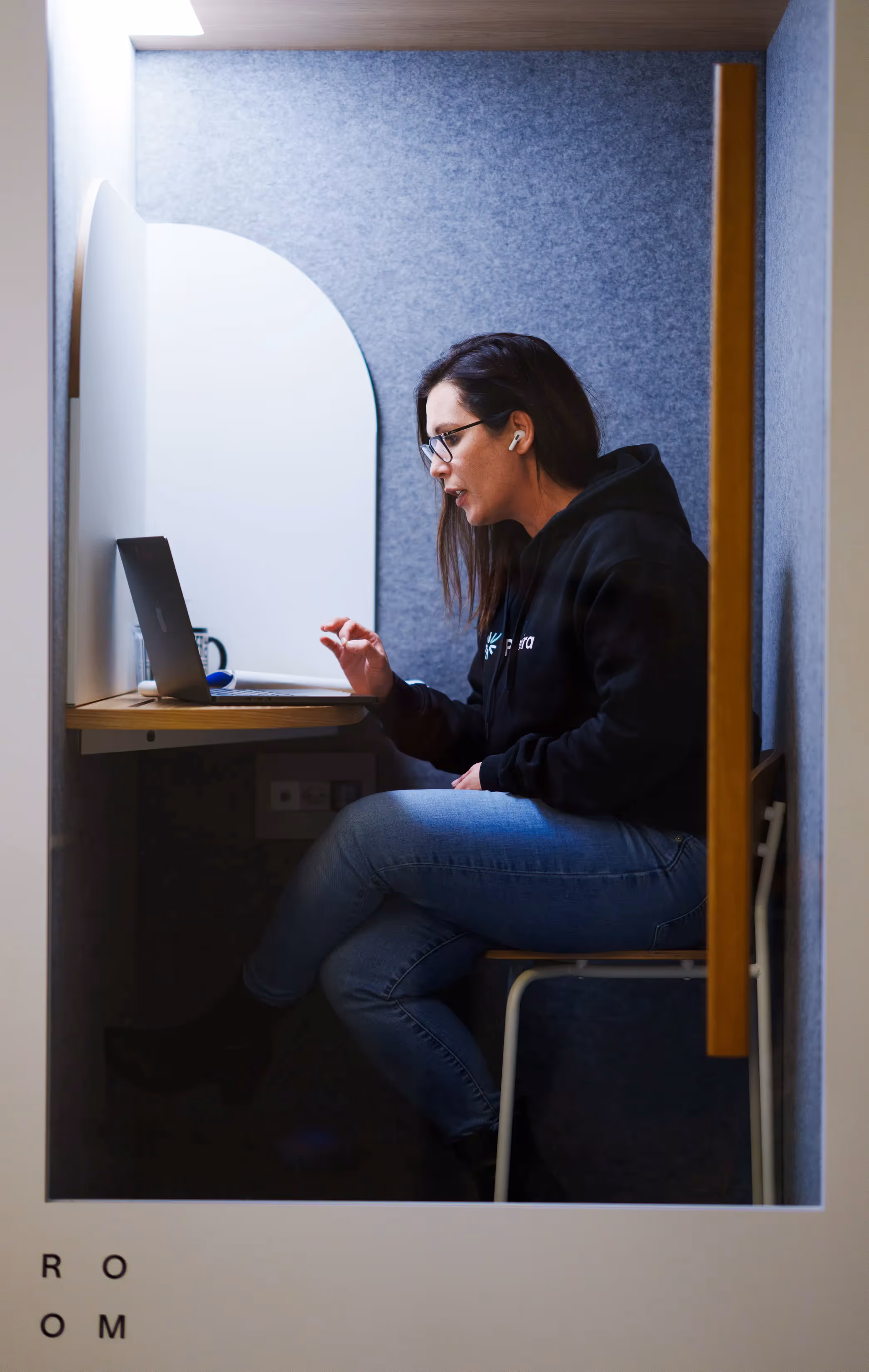 Woman wearing glasses and a black hoodie sits in a small booth, using a laptop and wearing wireless headphones.