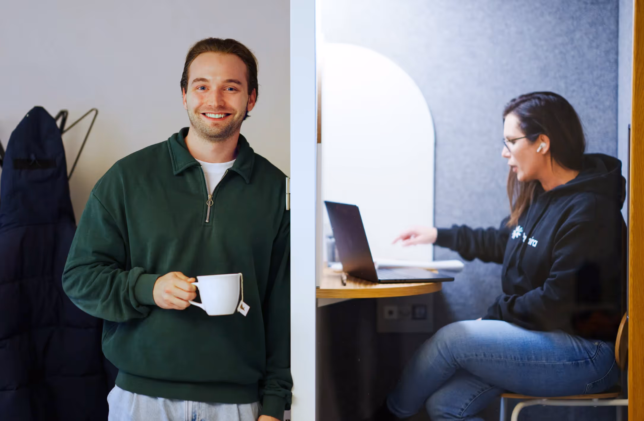 Smiling man in a green sweater holding a cup of tea, with a woman on a laptop in the background.