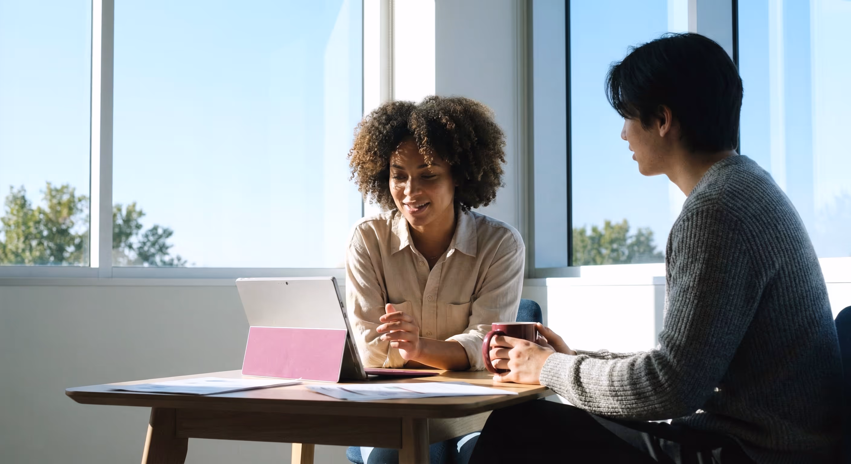 Two people are sitting at a table in daylight, looking at a tablet; one is holding a cup.