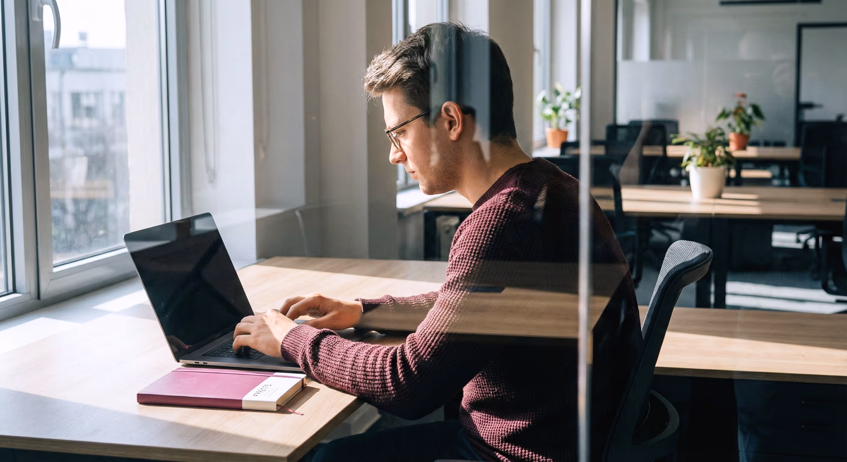 A man wearing glasses and a dark red sweater is working intently on a laptop at a bright desk with window light.