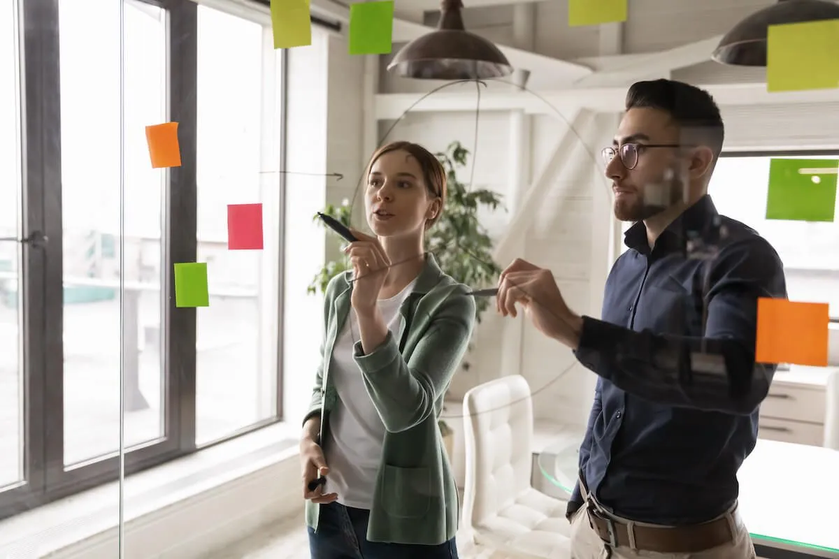 Two colleagues collaborating in an office, writing on a glass wall covered with colorful sticky notes during a brainstorming or planning session.