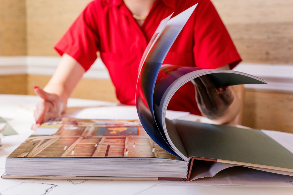 Person in a red shirt flipping through pages of a large book on a table.