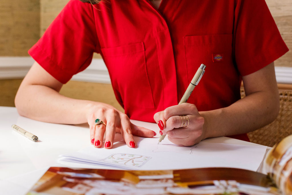 Person in a red shirt drawing on white paper with a beige pen, showing hands with red nail polish and rings.