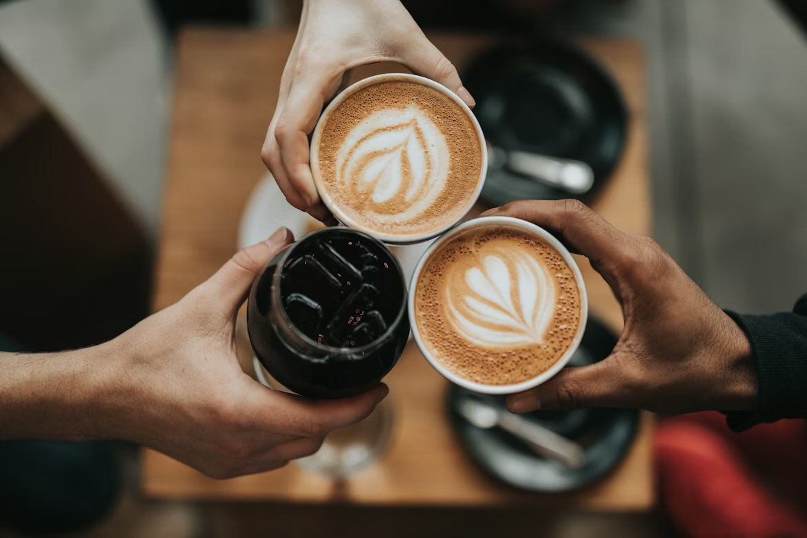 Trois tasses de café qui se touchent. Deux sont des lattes dans des tasses en céramique blanche. L’autre est un café noir glacé dans un verre