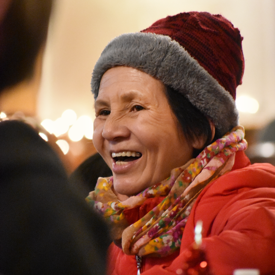 Smiling woman wearing a red jacket, floral scarf, and knitted hat at a festive indoor gathering.