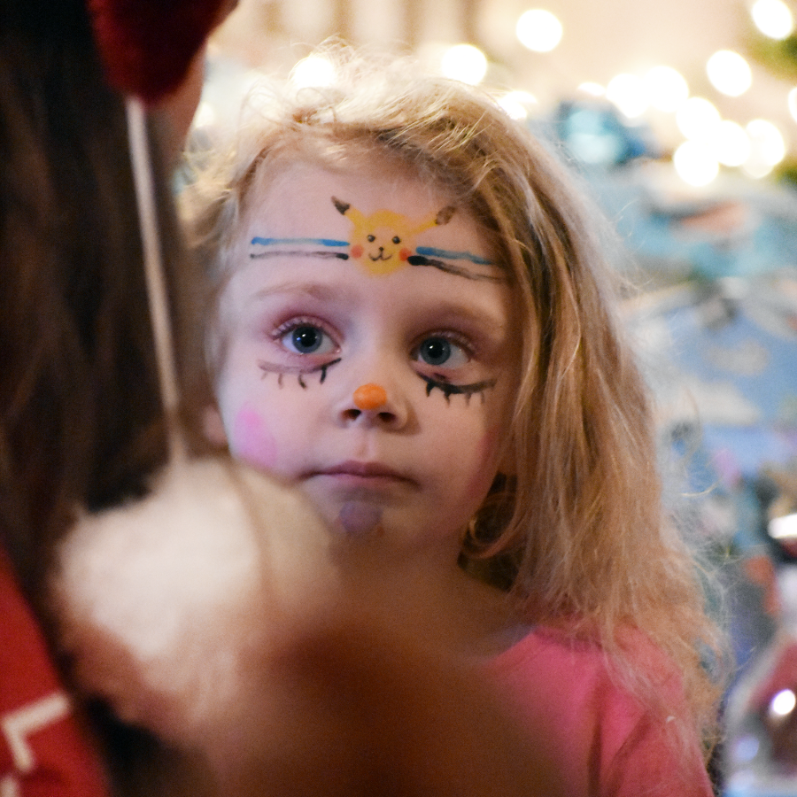 Young child with long blond hair and face paint including a yellow animal face on forehead and orange nose, looking slightly upward.