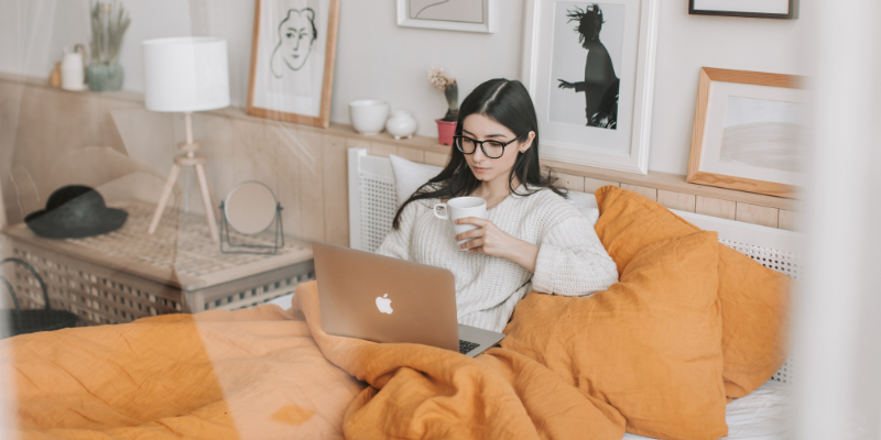 A woman in glasses sits on a bed with orange bedding, working on a laptop and holding a mug. The room has framed art and a cozy, relaxed vibe.