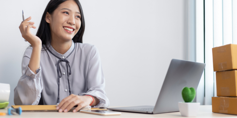 A woman in a light gray blouse smiles while holding a pen at a desk with a laptop, small plant, and stacked boxes, conveying a cheerful, productive mood.