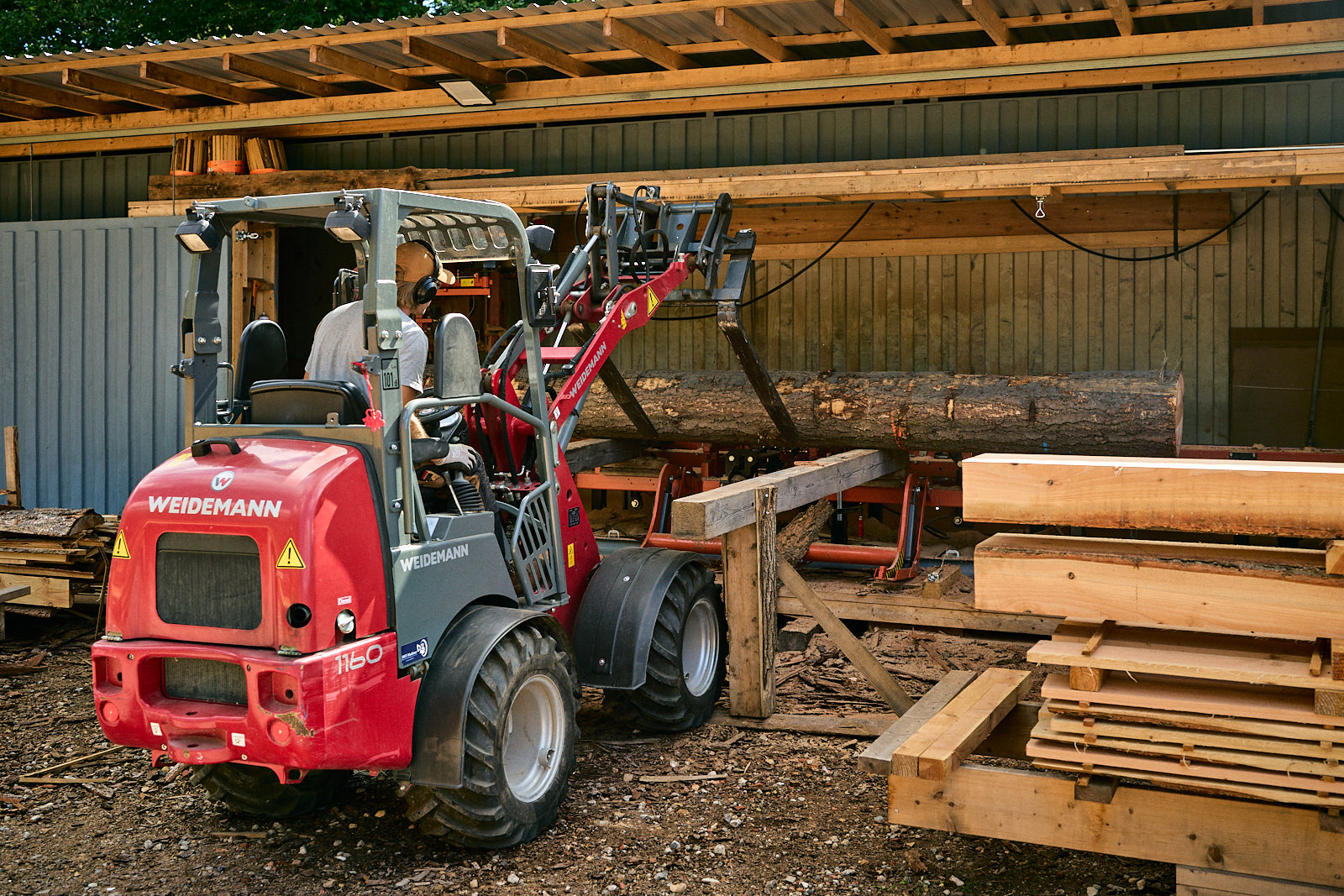 Man operating red Weidemann loader lifting a large log inside a wooden shed with stacked lumber nearby.