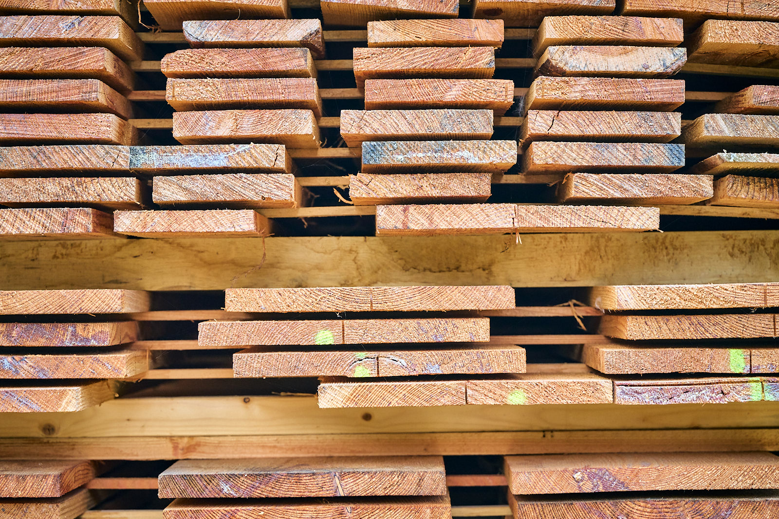 Stacked wooden planks with visible grain and some green markings stored in a lumberyard.