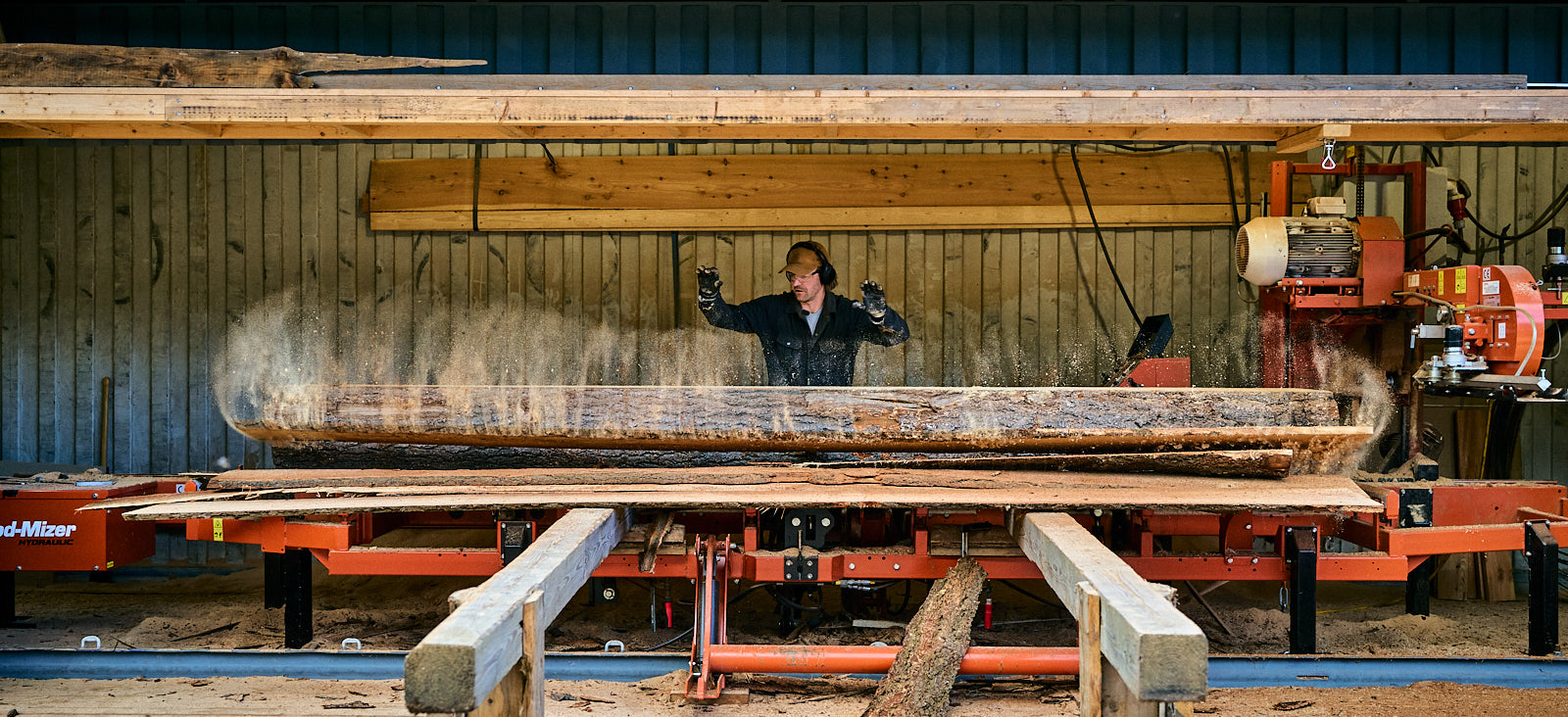 Man wearing protective ear muffs operating a large sawmill machine cutting a log in a woodshop.
