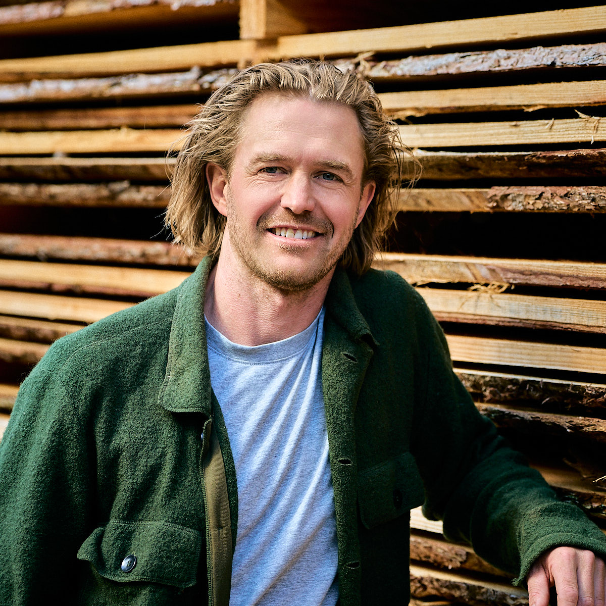 Smiling man with shoulder-length blonde hair wearing a green jacket and gray shirt, standing in front of stacked wooden planks.