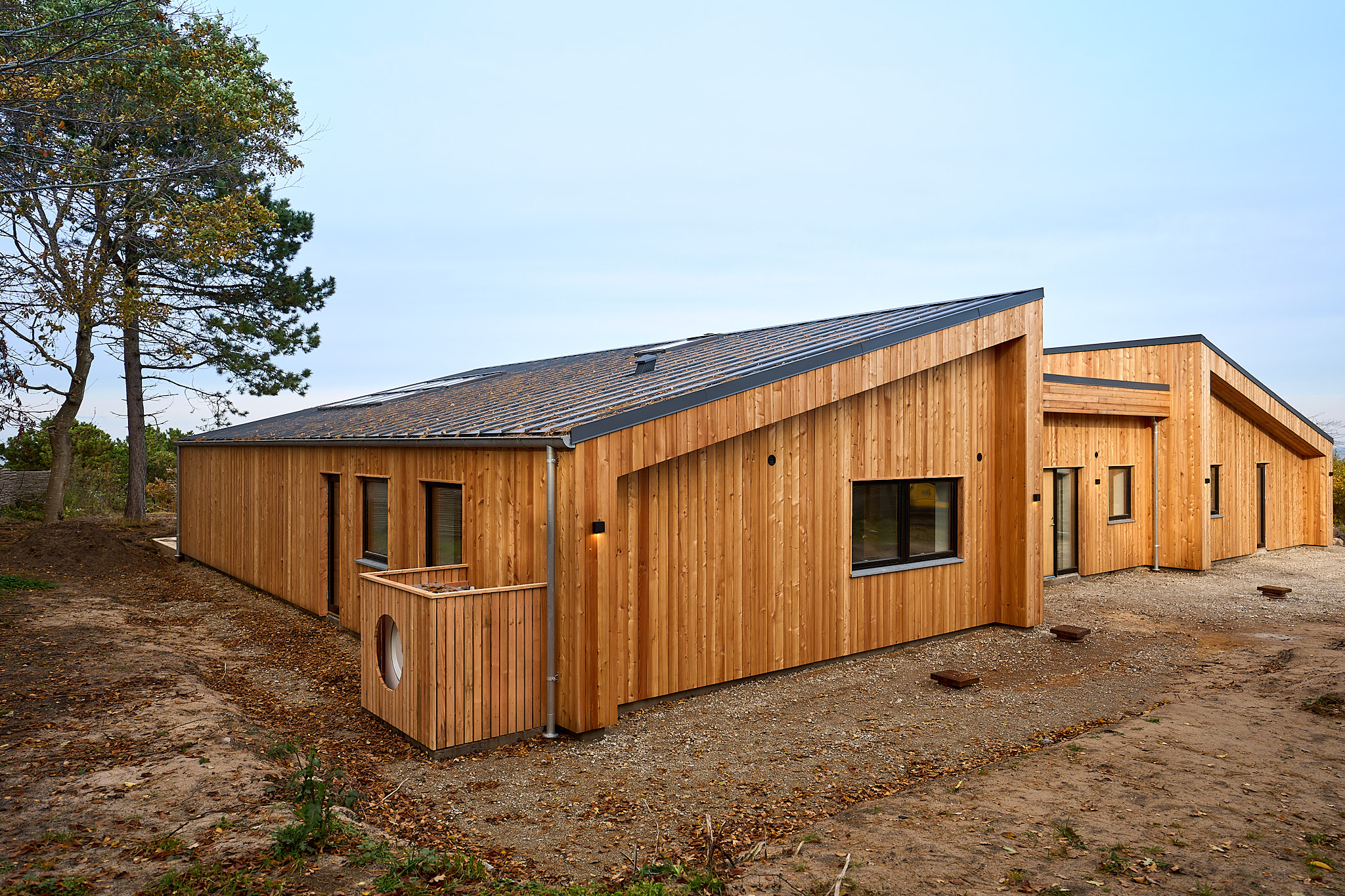 Modern single-story house with angled roofs and exterior clad in vertical wooden panels surrounded by bare soil and sparse vegetation.