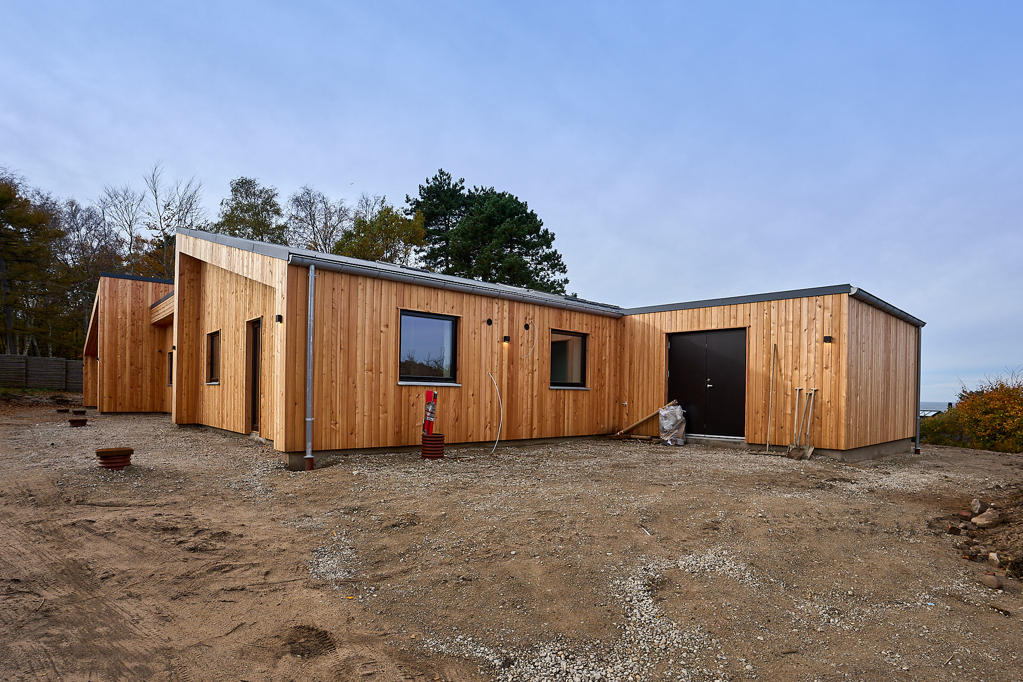 Modern single-story wooden building with dark doors and windows on a gravel lot under a clear sky.