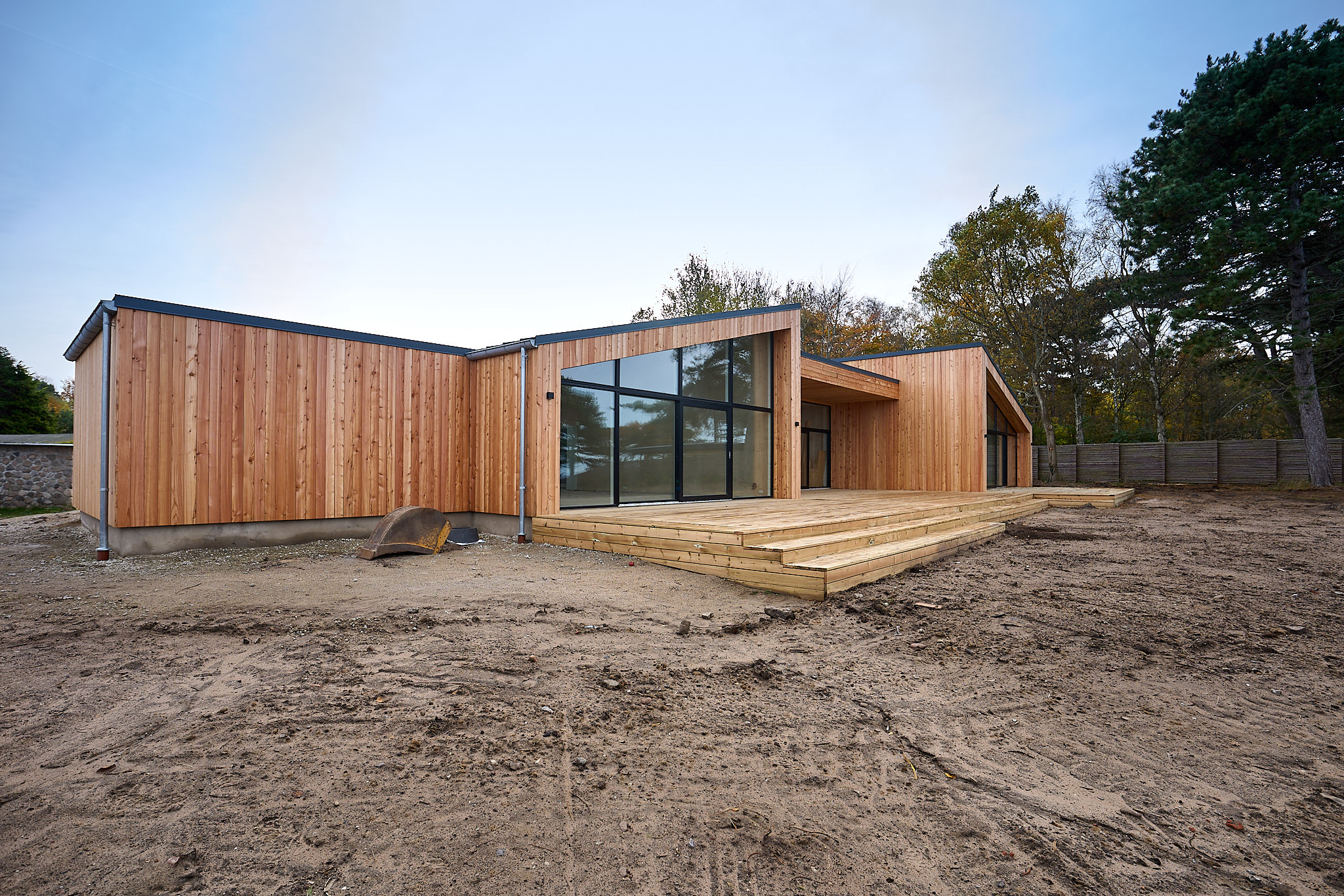 Modern single-story wooden house with large glass windows and a spacious wooden deck, set on a bare dirt yard.