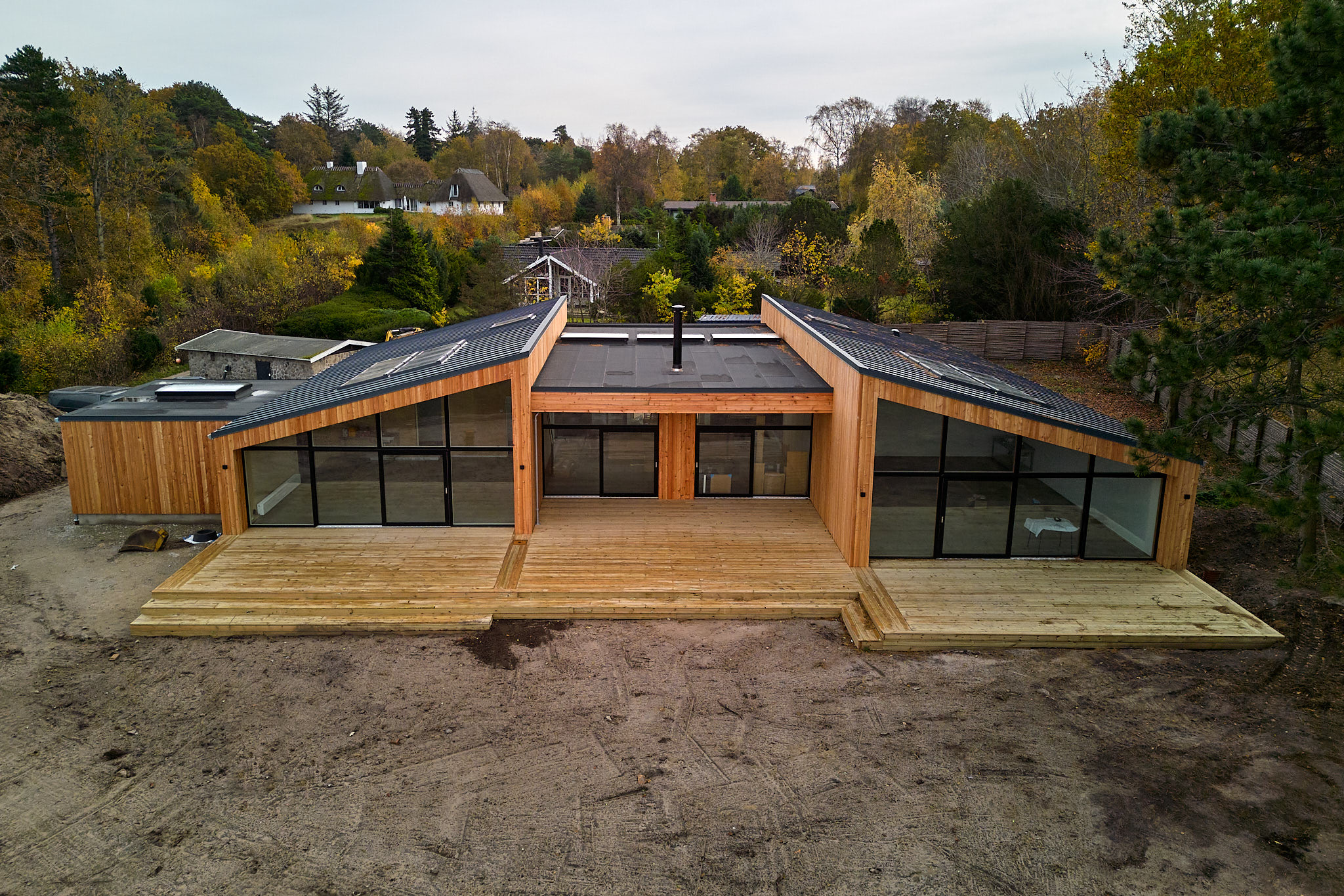 Modern wooden house with large glass windows and two slanted roofs, surrounded by trees in autumn.