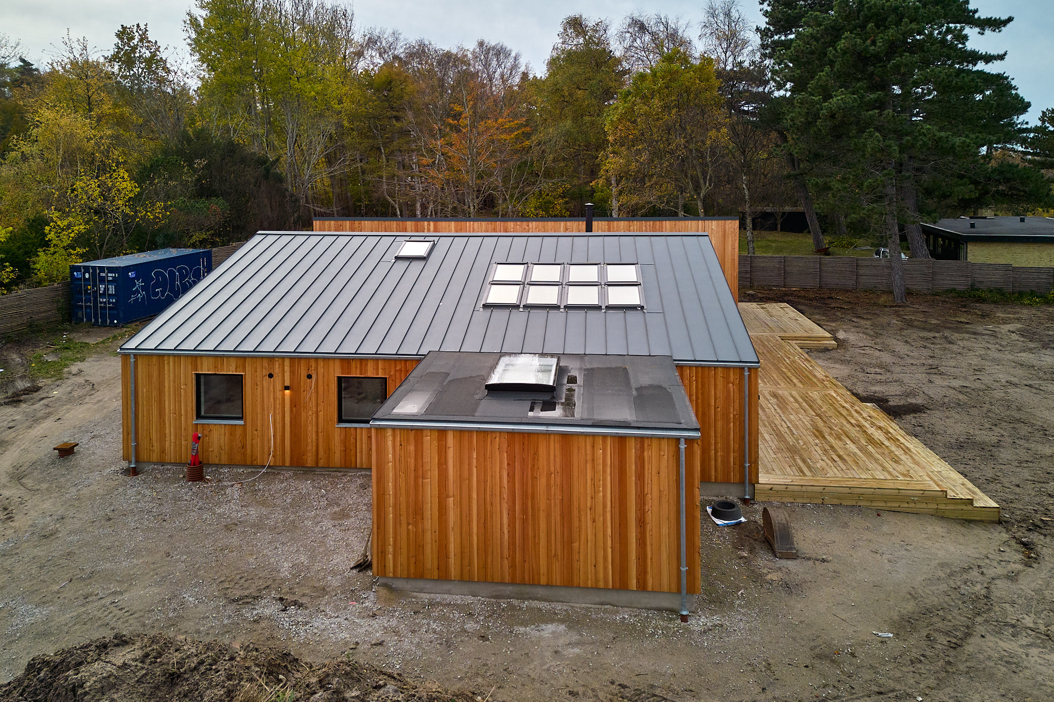 Modern single-story wooden house with a metal roof featuring multiple skylights, surrounded by dirt and trees.