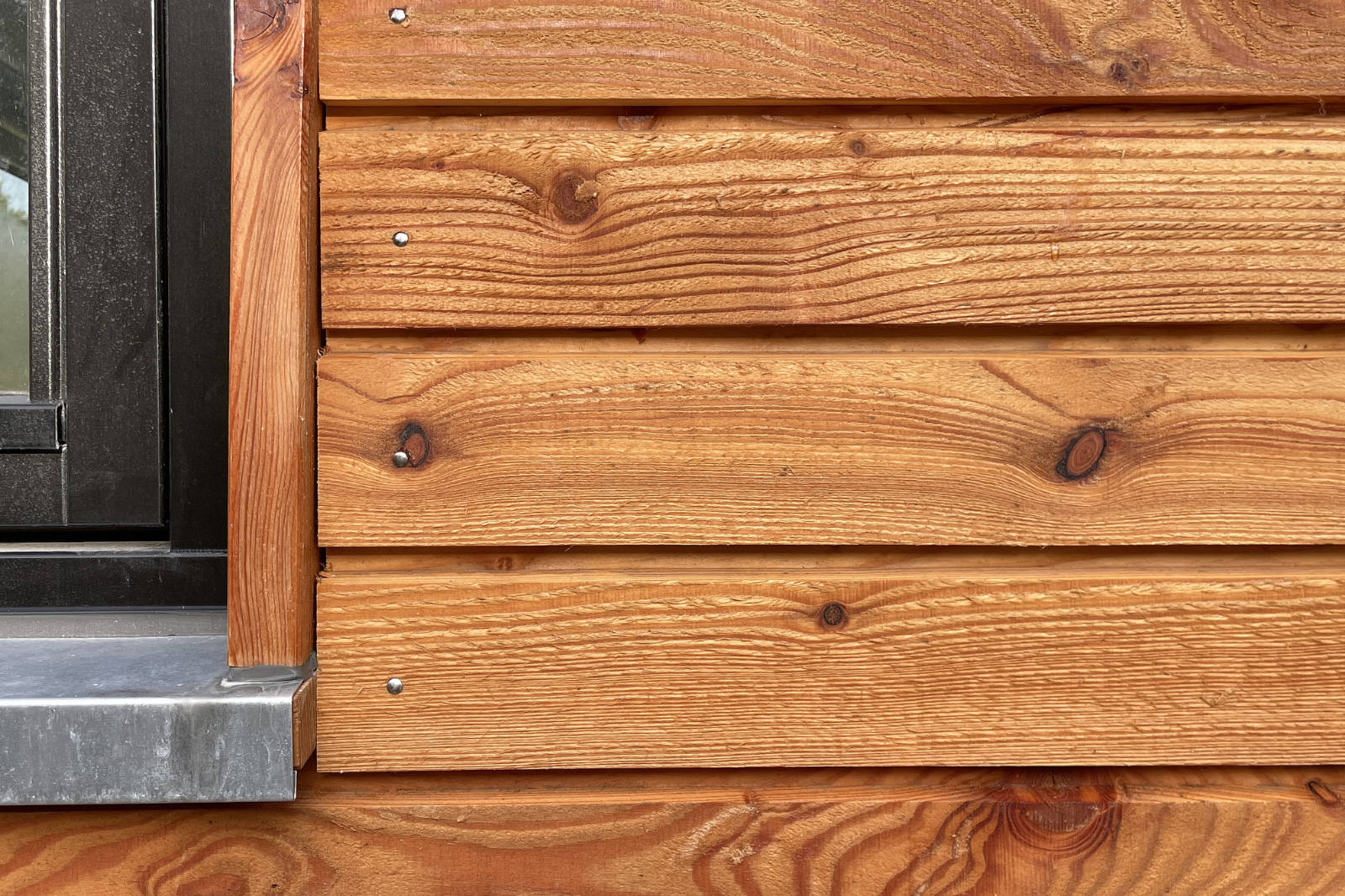 Close-up of a wooden exterior wall paneling next to a black window frame with a metal windowsill.