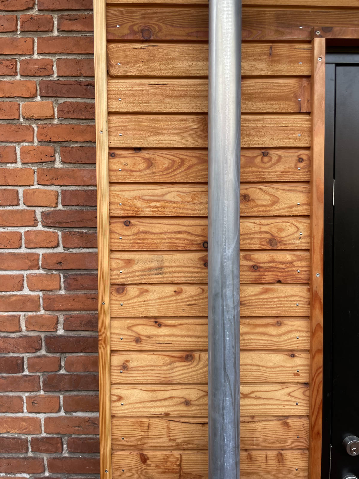 Close-up of a wall showing red brick on the left, horizontal wooden planks in the center, a vertical metal pipe, and a black door edge with handle on the right.