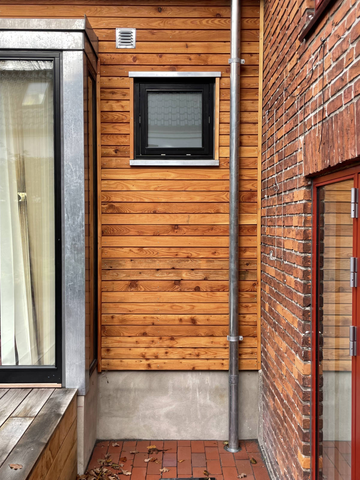 Exterior corner of a building showing a small black-framed window on wooden siding, a metal drainpipe, red brick wall, glass door with red trim, and red brick floor with scattered dry leaves.