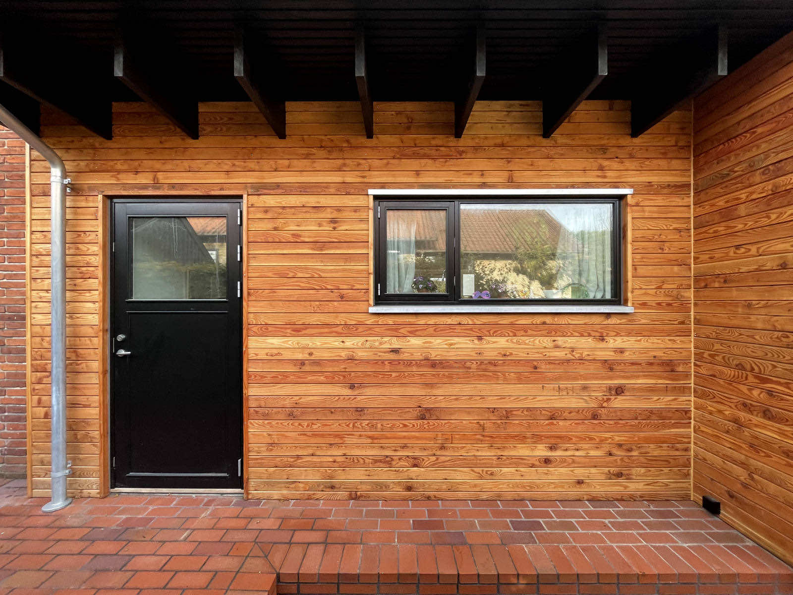 Wood-paneled exterior wall with a black door and a window reflecting a tiled roof and greenery.