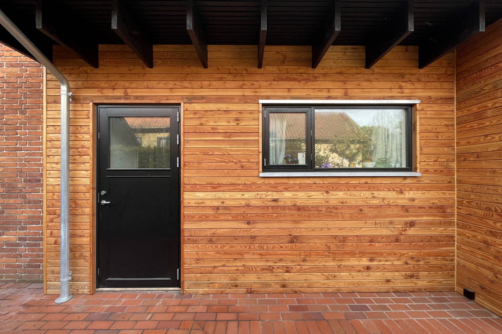 Exterior view of a wooden wall with a black door and a window with reflections of nearby houses and plants inside.