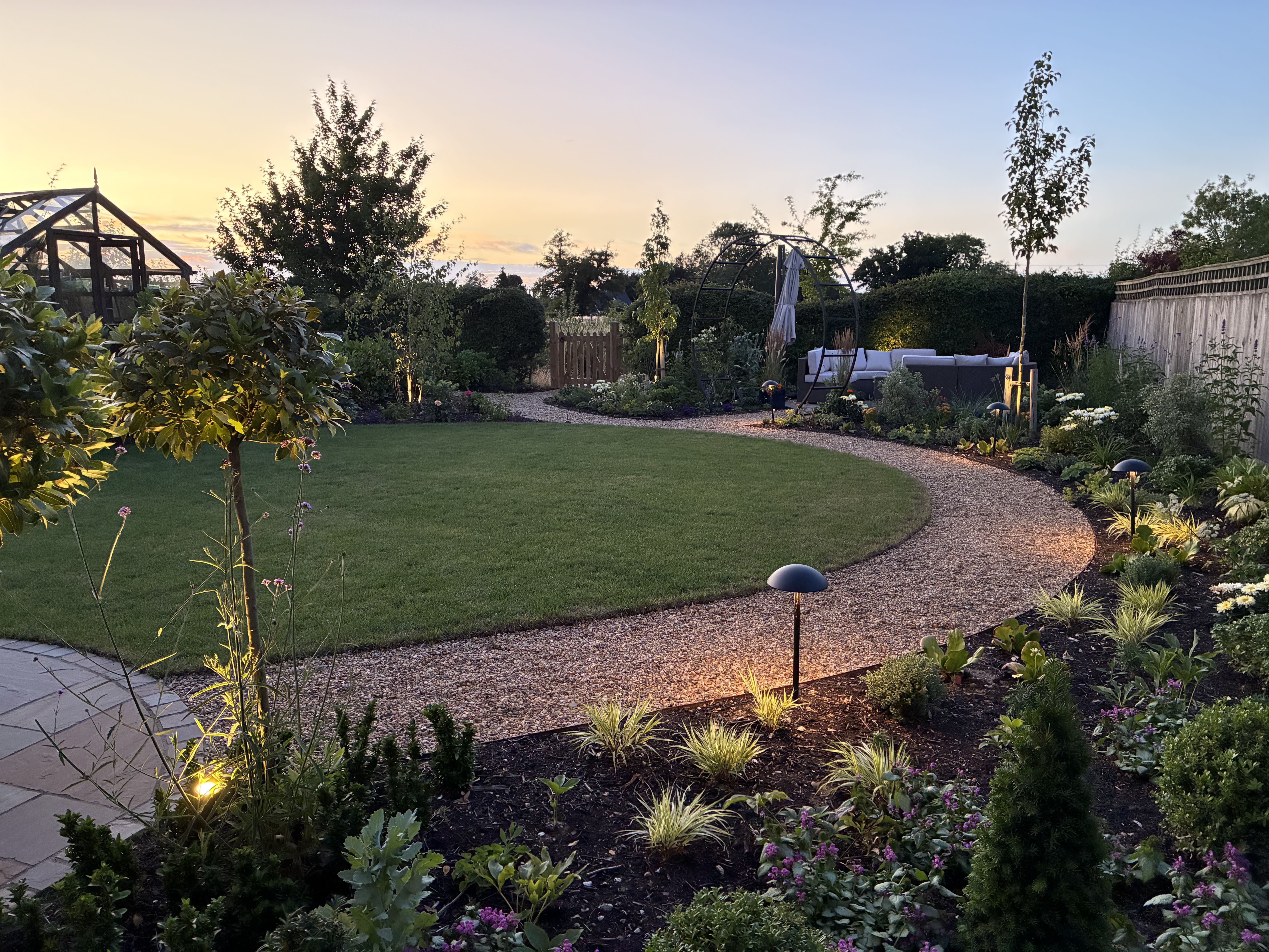 Curved gravel pathway winding round a luscious green lawn on one side and following a flower bed on the other. the picture is taken as the sun is setting and there warm mood lighting coming from between the plants in the flower bed.