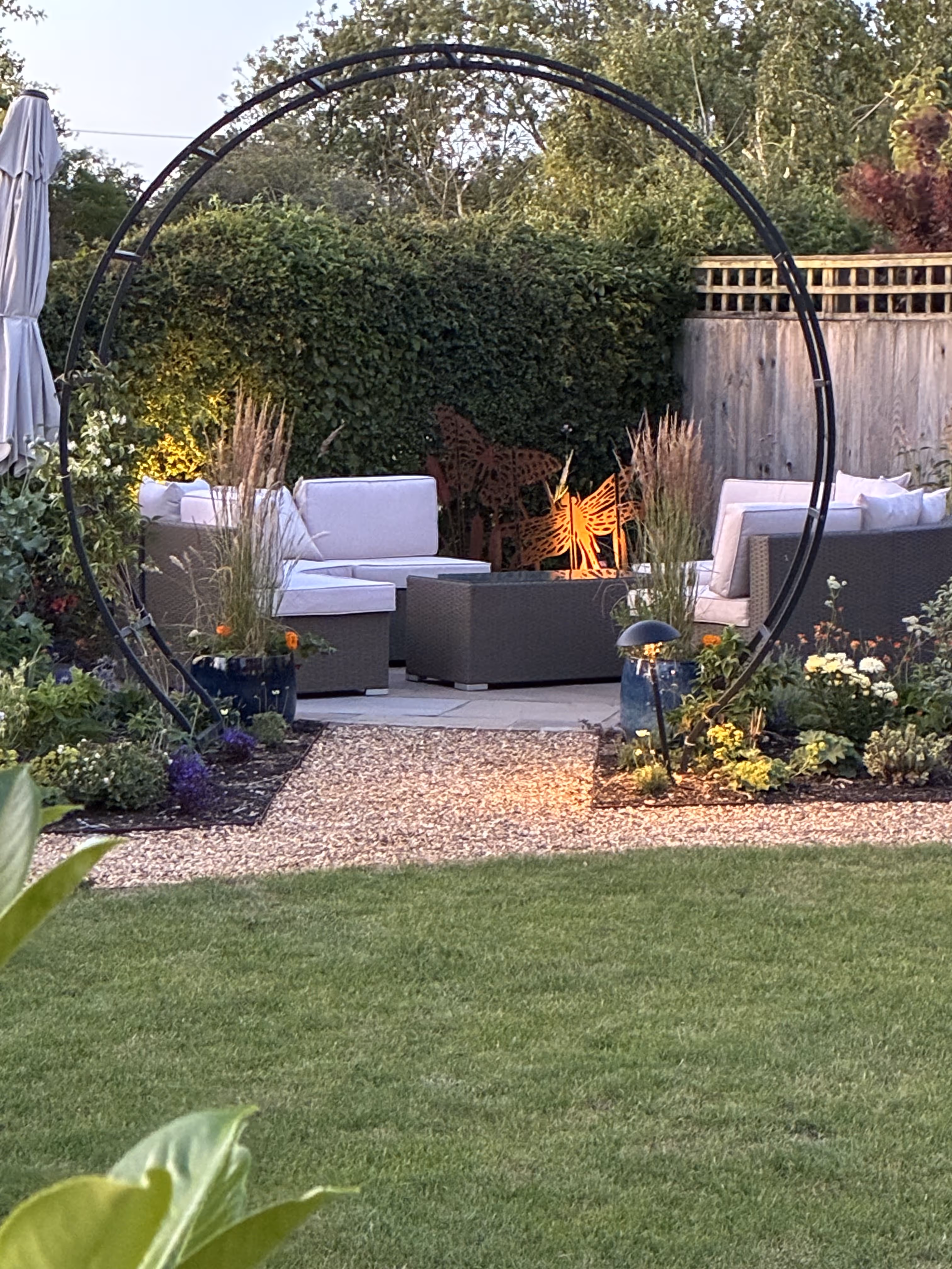 A cosy patio area in corner of garden with white outdoor furniture sitting on the paved slabs. The area is partially secluded by flowers and other tall plants with an circular black iron pergola arching over the entrance to the area. The picture is taken from further down the garden which shows the curved gravel pathway wrapping round the lawn leading to the cosy patio area.