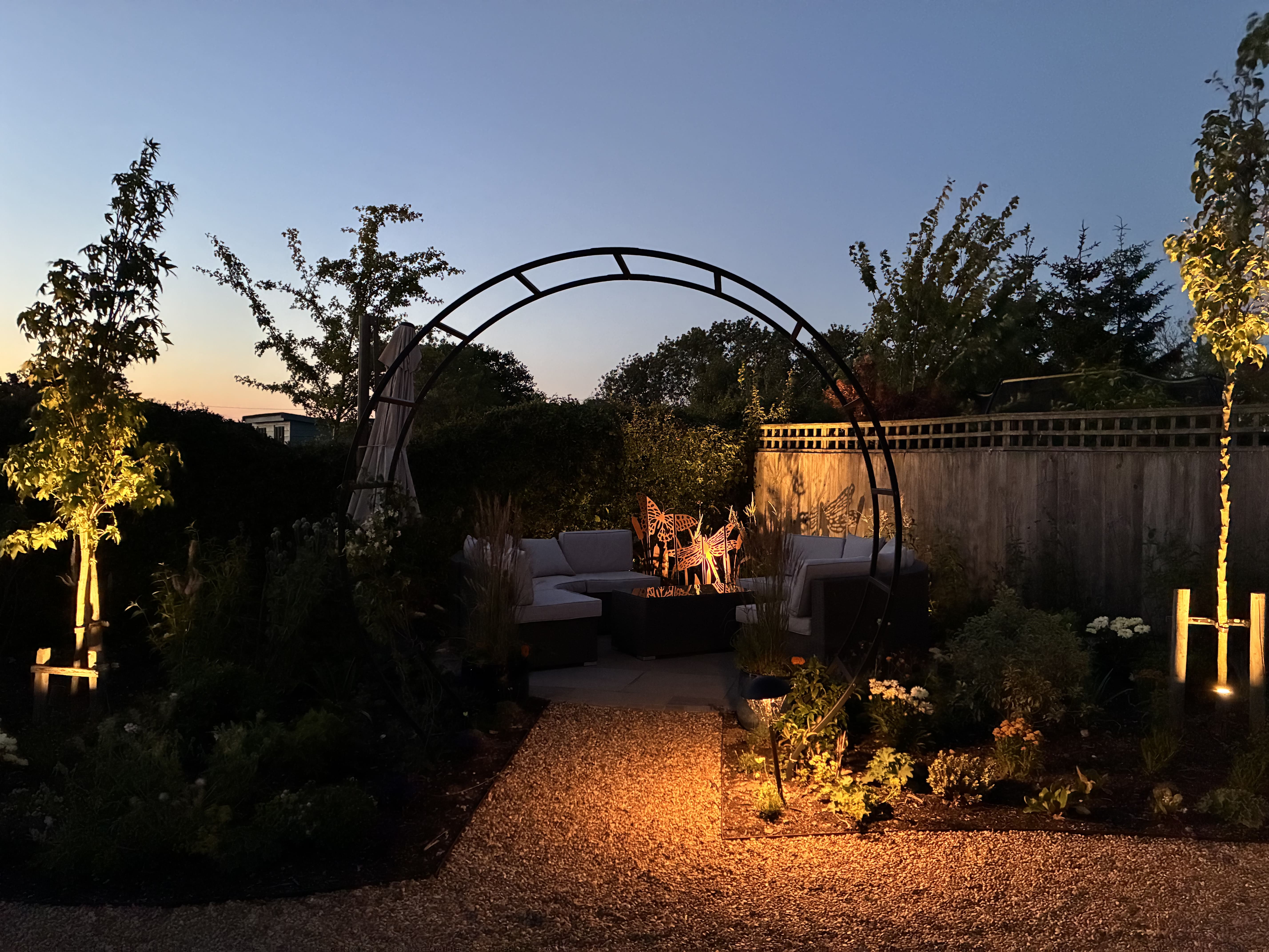 A cosy patio area in corner of garden with white outdoor furniture sitting on the paved slabs. The area is partially secluded by flowers and other tall plants with an circular black iron pergola arching over the entrance to the area. The sun is setting but the pathway leading to the area and the patio area itself is surrounded by warm lighting protruding from the greenery and vegetation surrounding the area. 