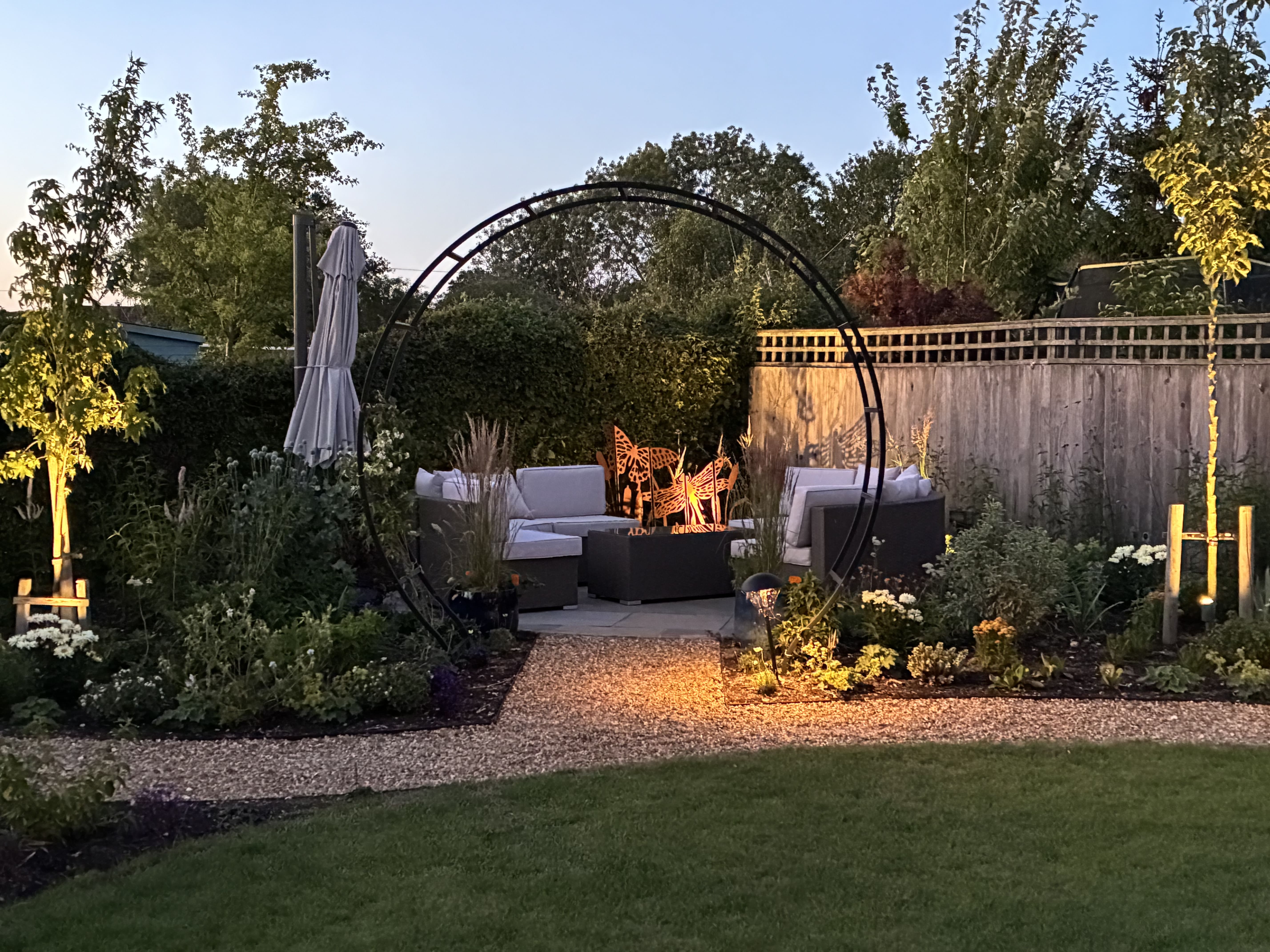 A cosy patio area in corner of garden with white outdoor furniture sitting on the paved slabs. The area is partially secluded by flowers and other tall plants with an circular black iron pergola arching over the entrance to the area. The picture is taken from further down the garden which shows the curved gravel pathway wrapping round the lawn leading to the cosy patio area.