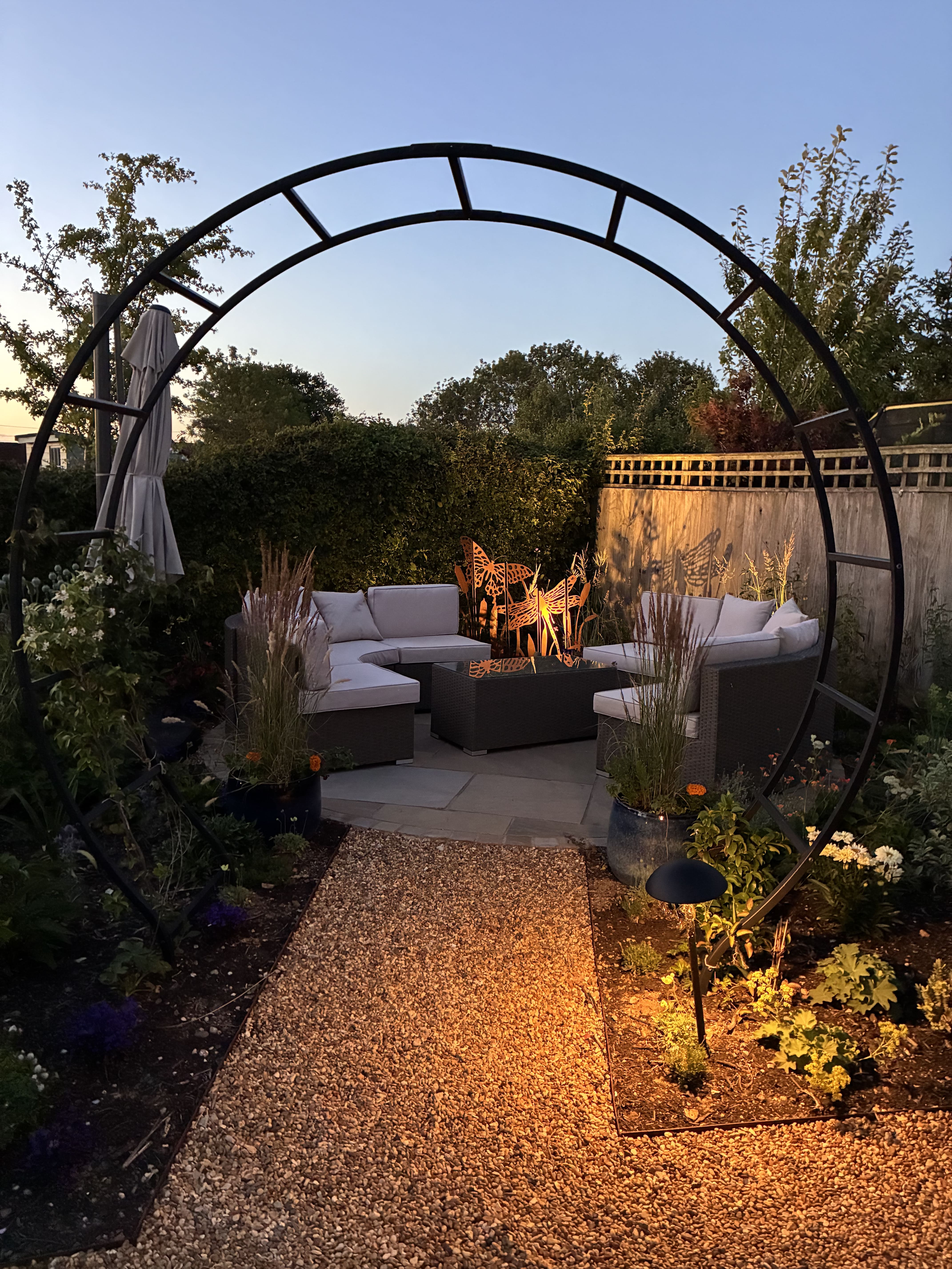 A cosy patio area in corner of garden with white outdoor furniture sitting on the paved slabs. The area is partially secluded by flowers and other tall plants with an circular black iron pergola arching over the entrance to the area.