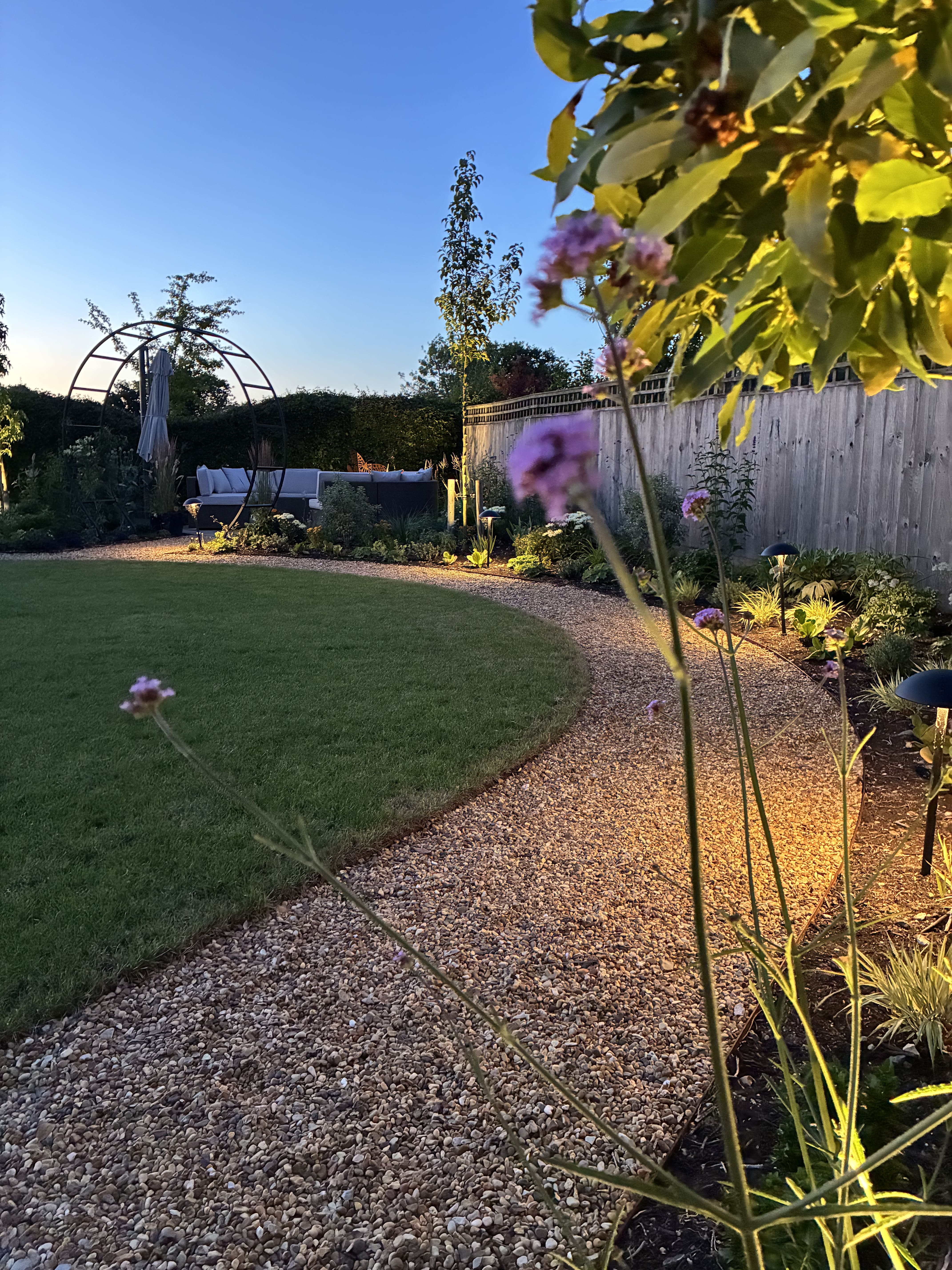 Curved gravel pathway winding round a luscious green lawn on one side and following a flower bed on the other. the picture is taken as the sun is setting and there warm mood lighting coming from between the plants in the flower bed.