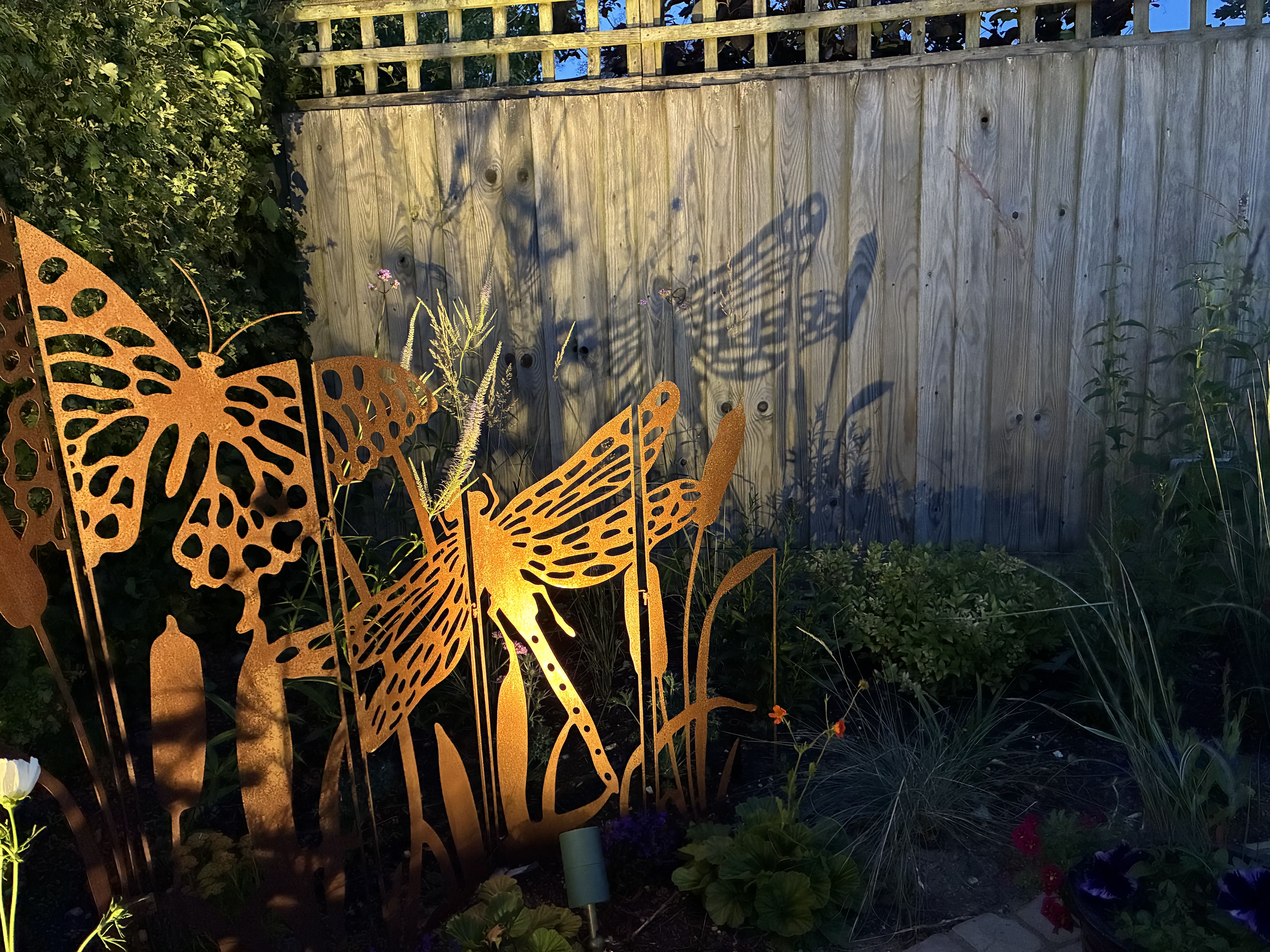 Decorative metal butterflies in corner of flower bed lit by warm ambient mood lighting at night.