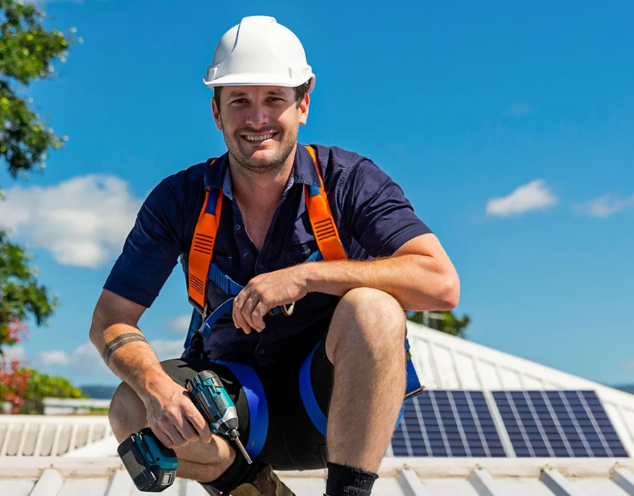 A worker in a hard hat and safety harness kneels on a roof holding a power drill, with solar panels visible nearby under a clear blue sky.