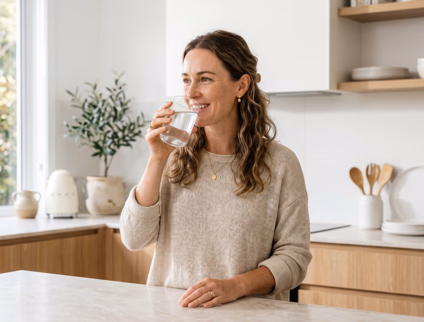 Smiling woman in beige sweater drinking a glass of water in a bright kitchen.